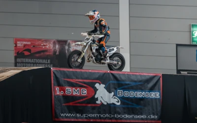 Fahrer in Schutzkleidung springt mit Motorrad &uuml;ber eine Rampe auf einer Messe.