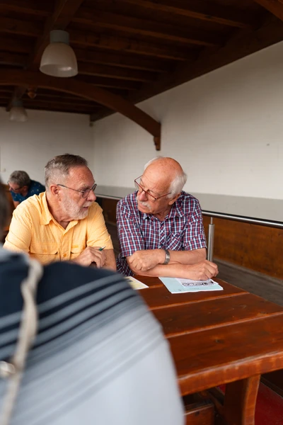 Zwei &auml;ltere M&auml;nner sitzen an einem Holztisch und unterhalten sich, Spa&szlig;rallye Bodensee 2026