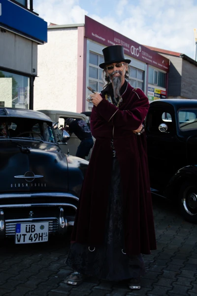 Person in eleganter Kleidung mit Zylinder vor klassischen Autos auf V8 Treffen