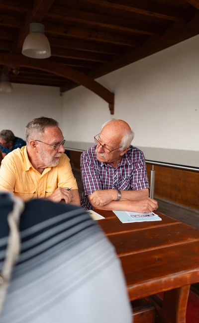 Zwei &auml;ltere M&auml;nner sitzen an einem Holztisch und unterhalten sich, Spa&szlig;rallye Bodensee 2026
